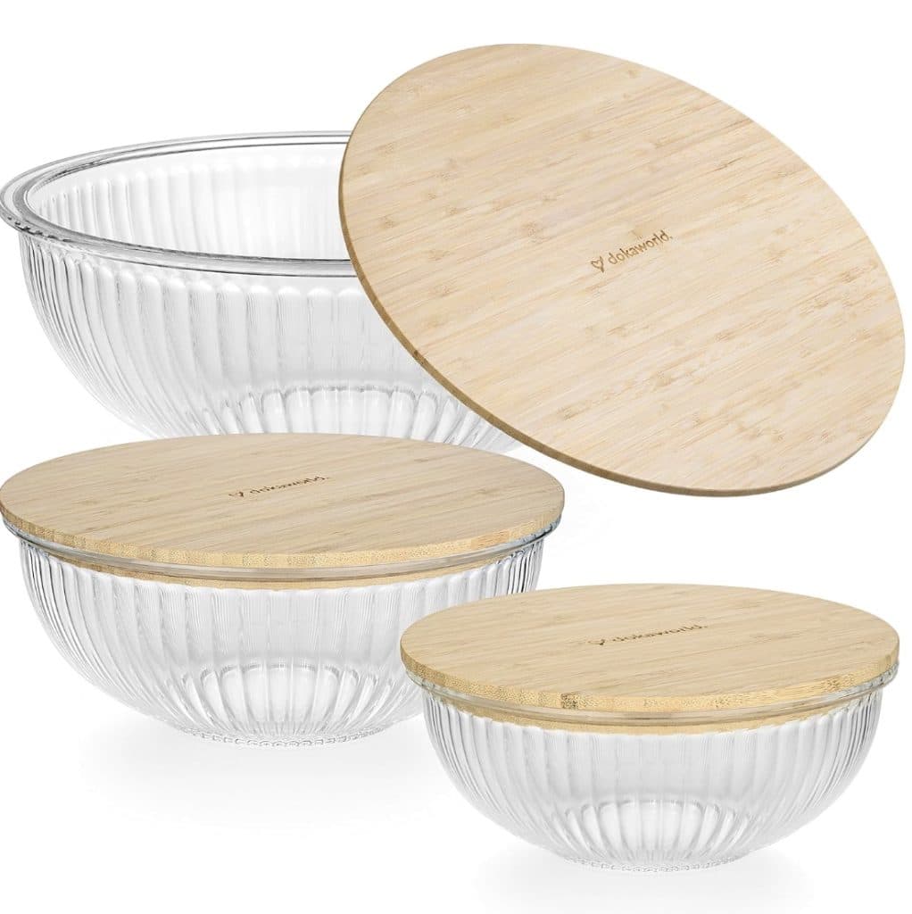 Three clear, ribbed glass bowls with wooden lids make a charming display for your favorite treats. Each bowl varies in size, with the largest on the top left and smallest on the bottom right. The bamboo lids add a natural touch, perfect for sharing Rice Krispie Treats on Valentine's Day.